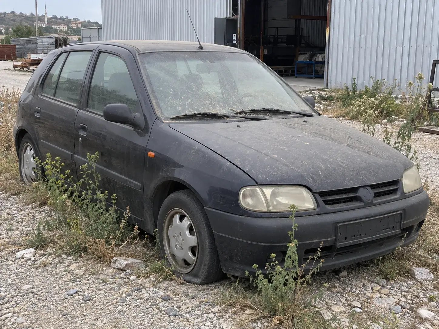 Ön lastiği inmiş, tozlanmış, plakası takılı olmayan koyu renkli hatchback; tekerleklerinin çevresinde yabani otlar büyümüş, sanayi sitesi görünümündeki bir sahada duruyor.