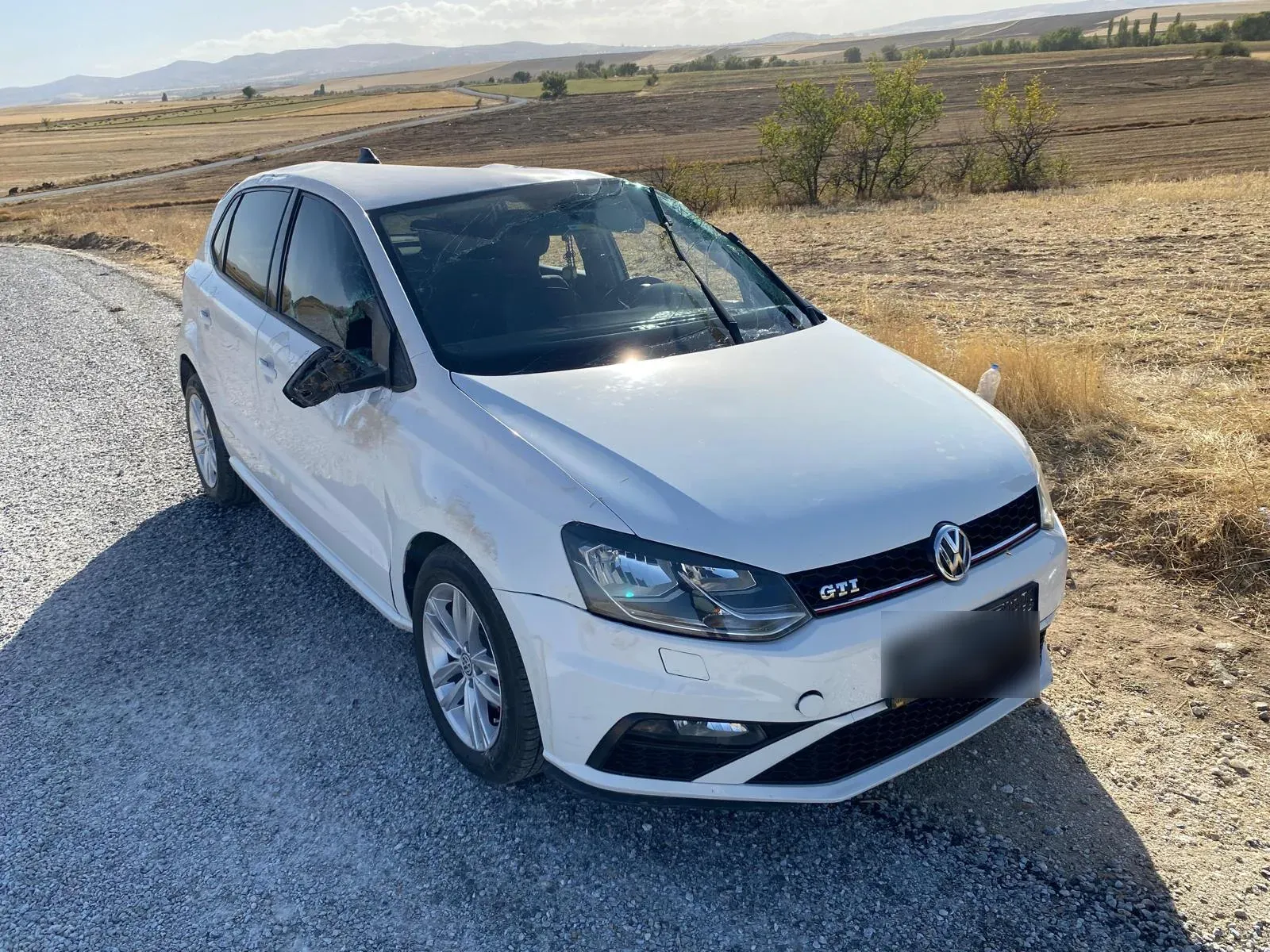 Yan paneli boydan boya ezilmiş, ön camı çatlamış ve yan aynası kırılmış beyaz Volkswagen Polo GTI, yol kenarında.