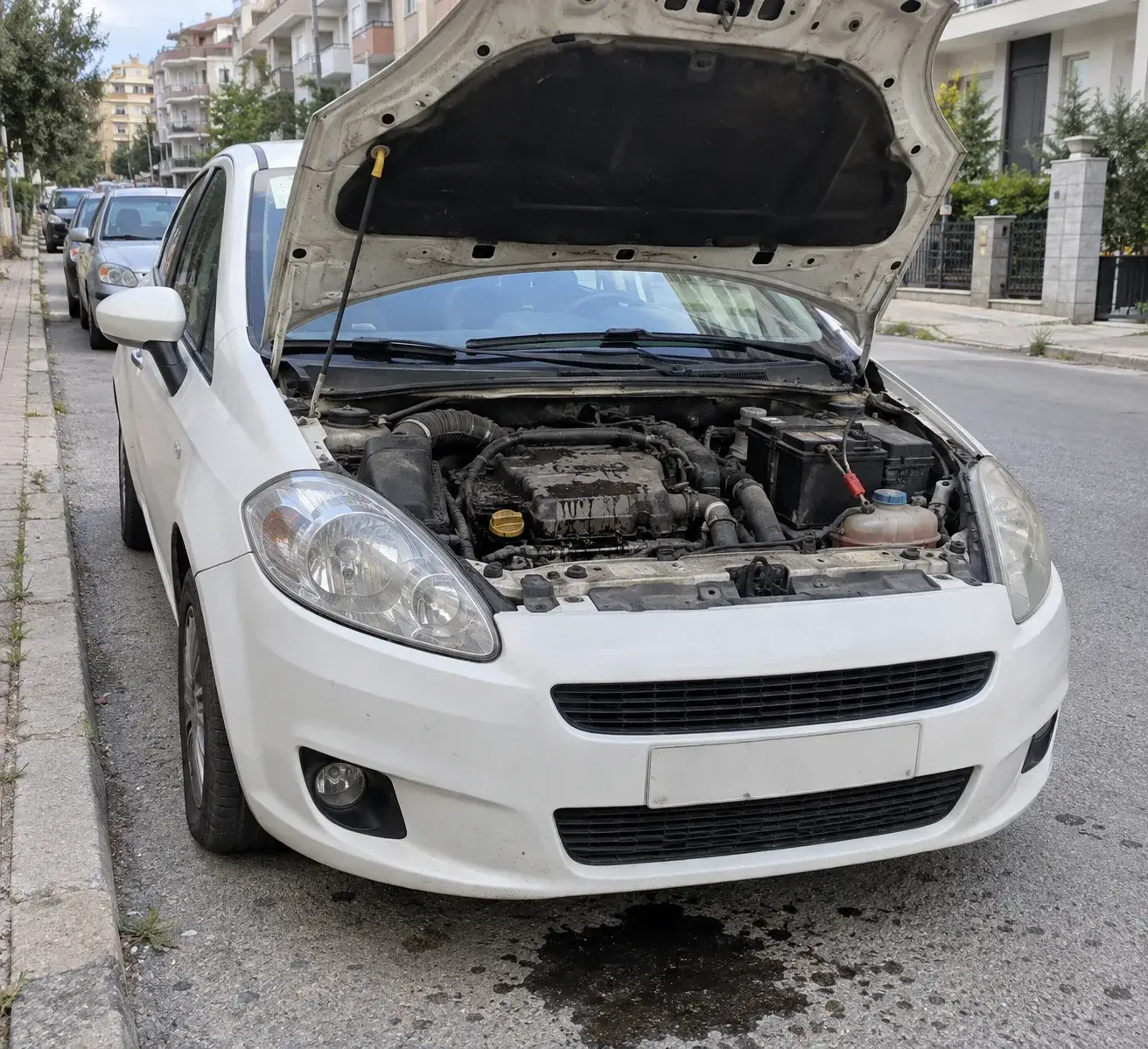Kaputu açık, motor bölümü yağ lekeli beyaz hatchback bir kaldırım kenarında park halinde; altındaki asfaltta koyu bir yağ birikintisi görünüyor.