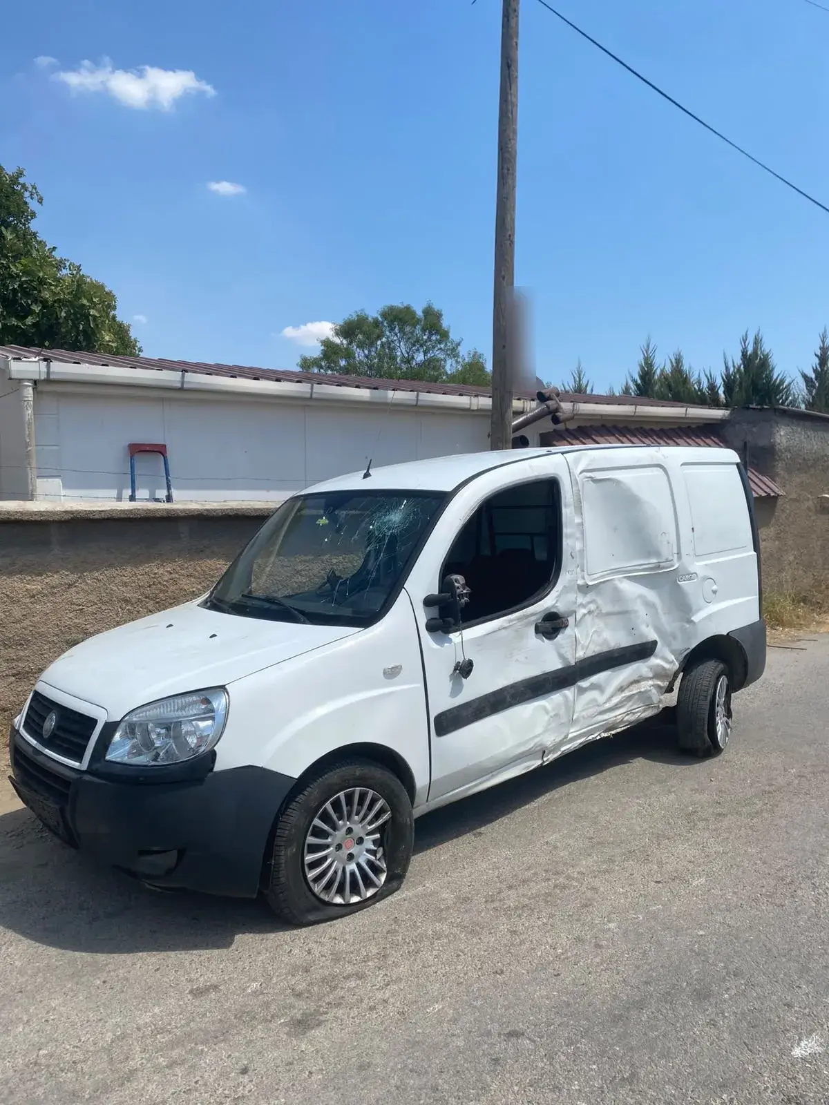 Yan paneli ve kapısı derin şekilde ezilmiş, ön camı çatlamış beyaz Fiat Doblo Cargo ticari araç.